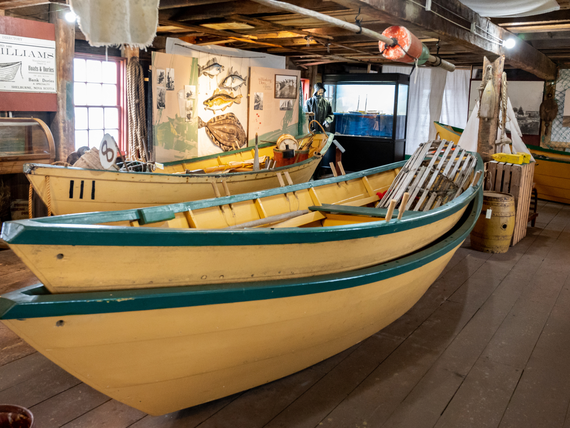 Two yellow Shelburne dory boats with green trim are stacked inside a museum space with wooden floors and exposed beams. Various exhibits and artifacts are visible in the background.