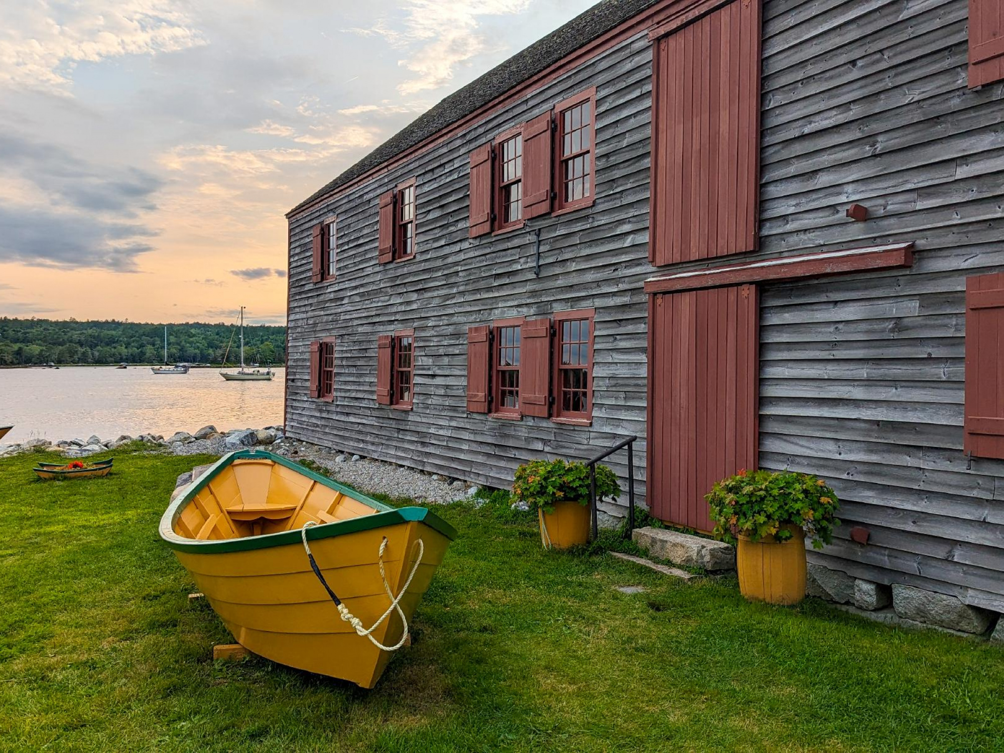 A bright yellow dory boat sits on green grass in front of a large, weathered gray wooden building with red trim. A body of water with sailboats is visible in the background under a cloudy sky.