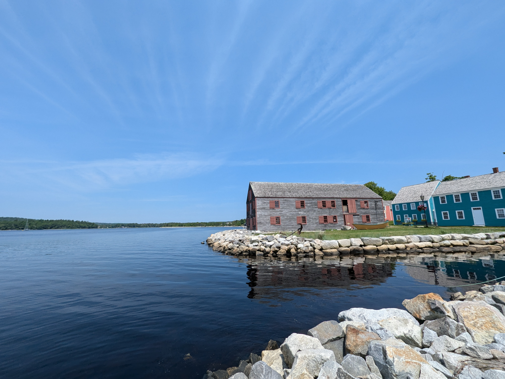 Two museum buildings, gray and blue, are on a rocky shore by water. The sky is blue with light clouds. Rocks are in the foreground.