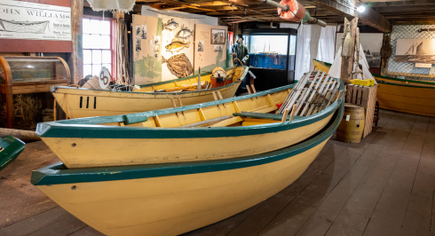 Two yellow Shelburne dory boats with green trim are stacked inside a museum space with wooden floors and exposed beams. Various exhibits and artifacts are visible in the background.