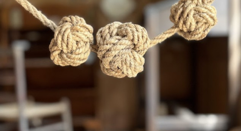Three tightly woven jute rope knots hang in a row, suspended by a thinner jute rope. The background is a blurred interior space, possibly a workshop or store, with shelves and a chair visible.